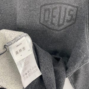 Pode incluir: Um moletom cinza escuro com a palavra "DEUS" impressa no canto superior direito. O moletom tem uma etiqueta com instru&ccedil;&otilde;es de lavagem e detalhes da composi&ccedil;&atilde;o do tecido. O tecido &eacute; 100% algod&atilde;o.