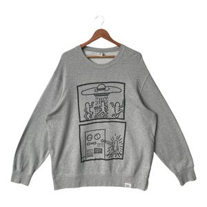 May include: A gray crewneck sweatshirt featuring a black graphic design. The design includes two rectangular panels with line drawings of figures, a UFO, and a control panel. The sweatshirt is hanging on a wooden hanger.