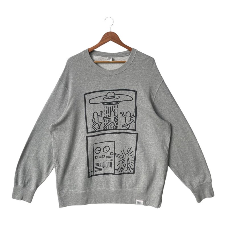 May include: A gray crewneck sweatshirt featuring a black graphic design. The design includes two rectangular panels with line drawings of figures, a UFO, and a control panel. The sweatshirt is hanging on a wooden hanger.