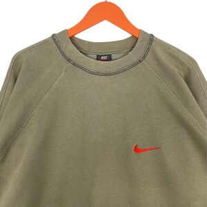 May include: Olive green Nike sweatshirt with a red Nike swoosh logo on the chest. The sweatshirt has a crew neck and raglan sleeves. The Nike logo is embroidered. The sweatshirt is hanging on an orange hanger.