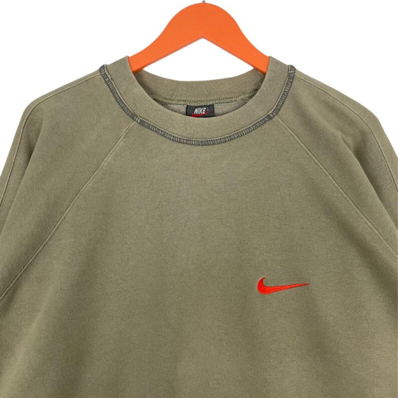 May include: Olive green Nike sweatshirt with a red Nike swoosh logo on the chest. The sweatshirt has a crew neck and raglan sleeves. The Nike logo is embroidered. The sweatshirt is hanging on an orange hanger.