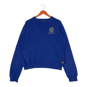 May include: A royal blue sweatshirt with a crew neck and long sleeves. A yellow cartoon character graphic is embroidered on the upper left chest. The sweatshirt has a small tag on the lower left sleeve.