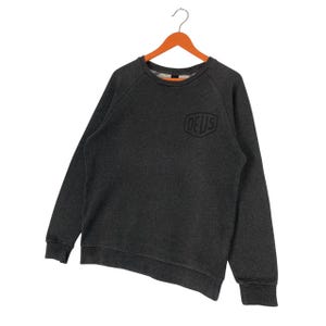 Pode incluir: Camisola cinza escuro com a palavra "DEUS" em fonte preta e estilizada no peito. A camisola tem mangas compridas e gola redonda. Est&aacute; pendurada num cabide de madeira laranja.