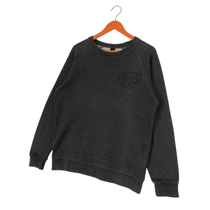 Pode incluir: Camisola cinza escuro com a palavra "DEUS" em fonte preta e estilizada no peito. A camisola tem mangas compridas e gola redonda. Est&aacute; pendurada num cabide de madeira laranja.