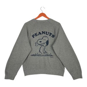 May include: A heather grey long-sleeve sweatshirt featuring a dark blue graphic print on the back. The print displays the word PEANUTS in an arched font above an illustration of the character Snoopy holding a bone in his mouth.
