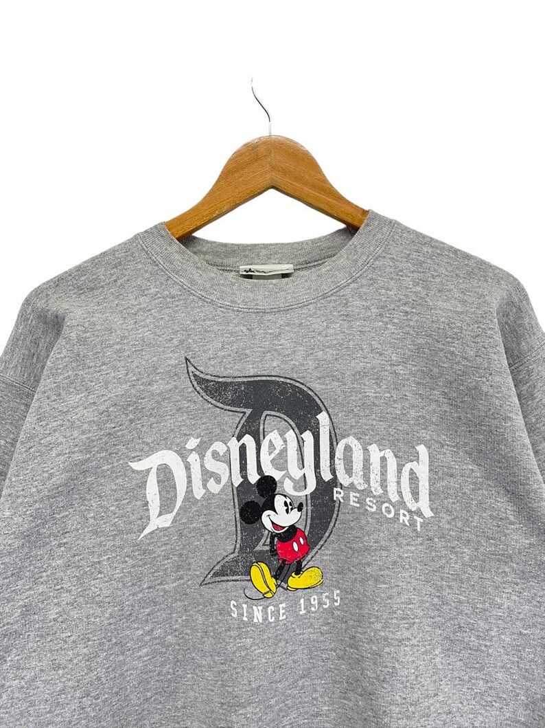 May include: A grey sweatshirt with the Disneyland Resort logo, Mickey Mouse, and the text "SINCE 1955." The graphic has a distressed, vintage appearance. The sweatshirt is displayed on a wooden hanger.
