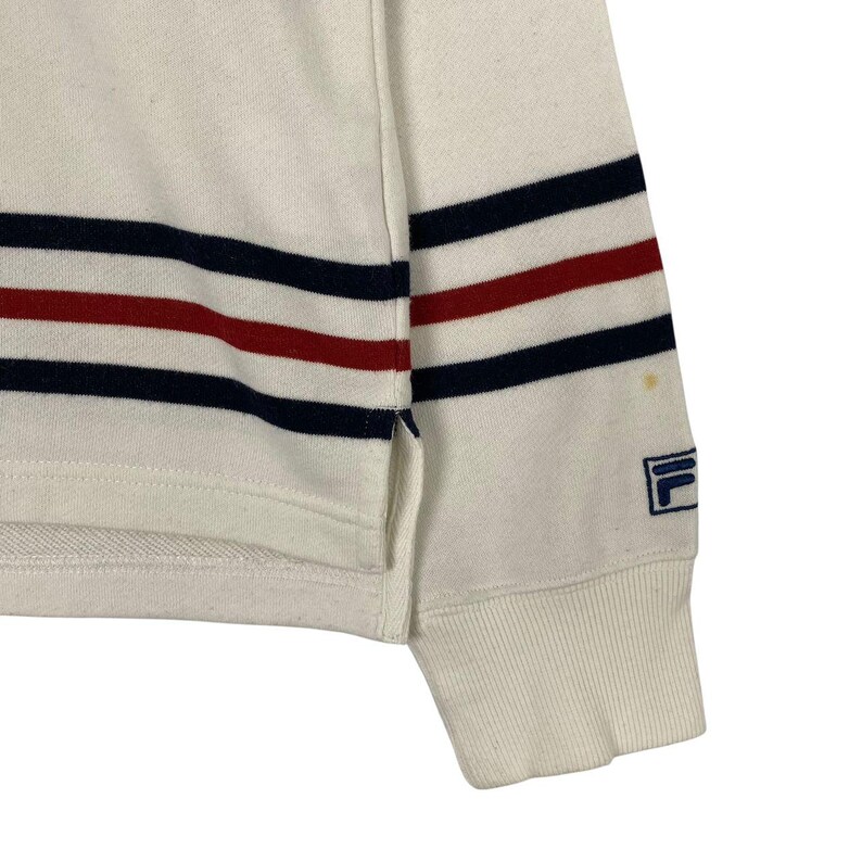 May include: A white long-sleeve sweatshirt featuring horizontal navy blue and red stripes across the torso and sleeves. The garment includes a side slit at the hem, ribbed cuffs, and a small blue Fila logo embroidered on the lower sleeve.