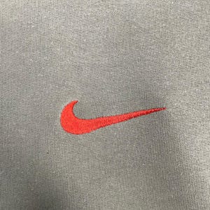 May include: Close-up of a gray fabric with a red Nike swoosh logo embroidered on the left side. The logo is a stylized check mark, a popular athletic brand symbol.