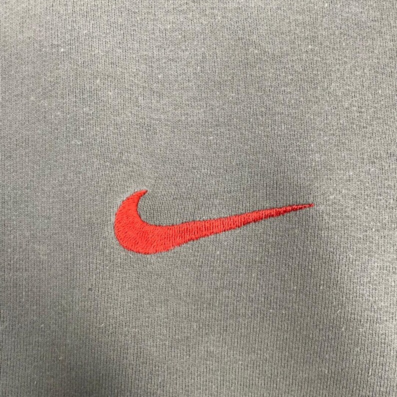 May include: Close-up of a gray fabric with a red Nike swoosh logo embroidered on the left side. The logo is a stylized check mark, a popular athletic brand symbol.