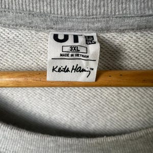 May include: Close-up of a gray sweatshirt with a ribbed neckline. A white tag displays the text "UT", "3XL", "MADE IN VIETNAM", and "Keith Haring&trade;". The sweatshirt is hanging on a wooden hanger.