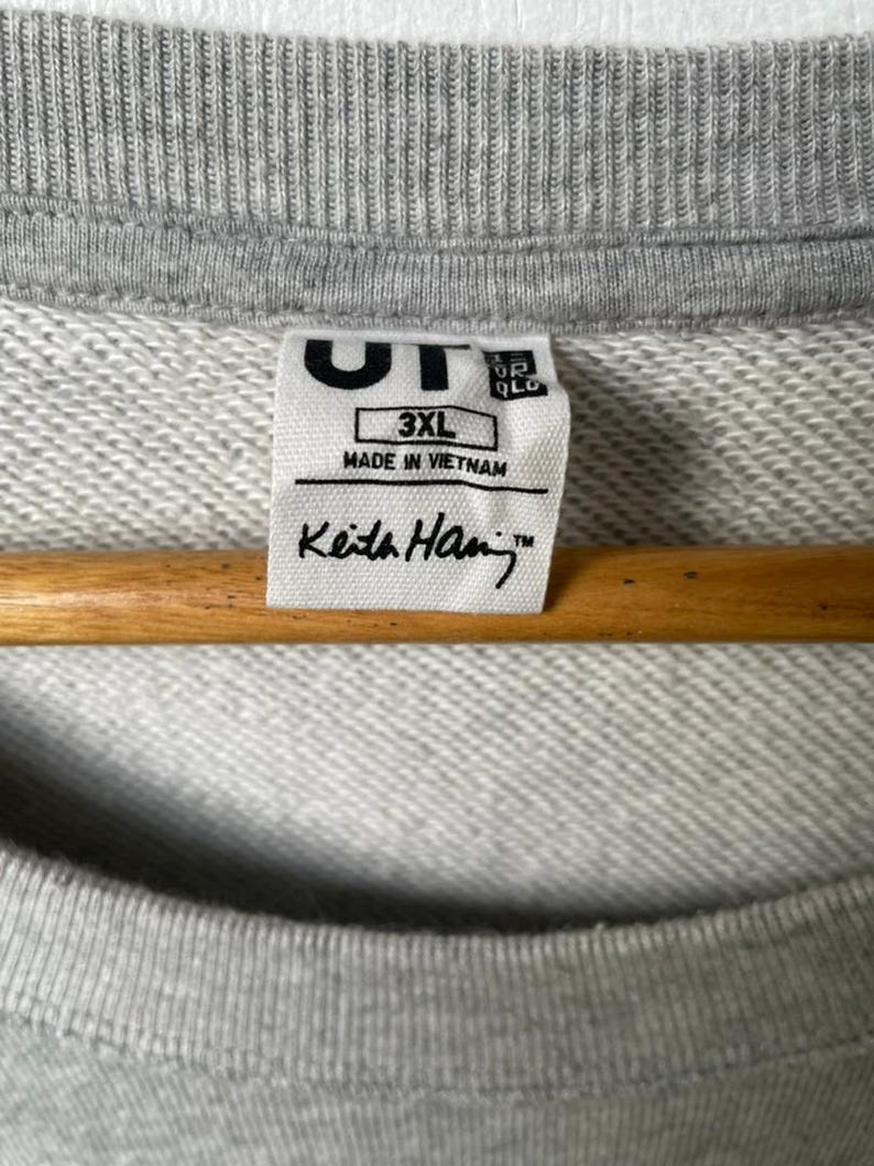 May include: Close-up of a gray sweatshirt with a ribbed neckline. A white tag displays the text "UT", "3XL", "MADE IN VIETNAM", and "Keith Haring&trade;". The sweatshirt is hanging on a wooden hanger.