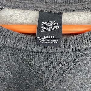 Pode incluir: Close-up de um moletom cinza com uma etiqueta preta. A etiqueta diz "Deus Ex Machina" e "SMALL", com "MADE IN CHINA" e "FABRIQUE EN CHINE" impressos abaixo. O moletom tem uma gola canelada e uma costura em forma de V.