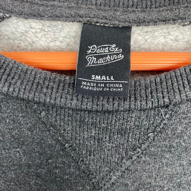 Pode incluir: Close-up de um moletom cinza com uma etiqueta preta. A etiqueta diz "Deus Ex Machina" e "SMALL", com "MADE IN CHINA" e "FABRIQUE EN CHINE" impressos abaixo. O moletom tem uma gola canelada e uma costura em forma de V.