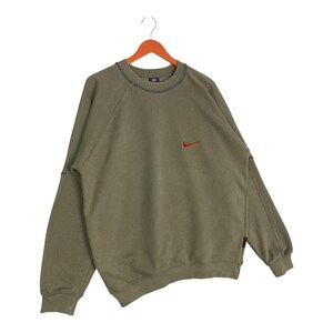 May include: Olive green sweatshirt with a small red Nike logo on the chest. The long-sleeved top has a crew neck and is hanging on a wooden hanger. The sweatshirt has a relaxed fit and a vintage look.