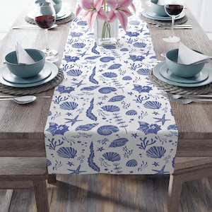 May include: A white table runner with a blue seashell pattern, starfish, and seaweed designs. The runner is placed on a wooden table set for a meal, with plates, bowls, and wine glasses. A vase of pink flowers is in the center.