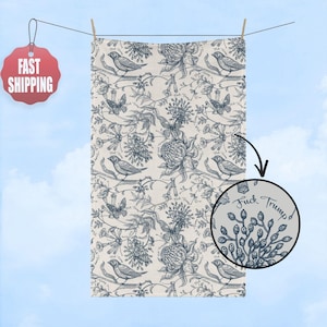 May include: A cream-colored towel with a blue floral and bird pattern, hanging on a clothesline. The design includes flowers, butterflies, and birds. A circular inset shows the text "Fuck Trump".