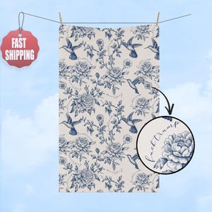 May include: A cream-colored towel with a blue floral and hummingbird pattern. The design includes large peony-like flowers and hummingbirds, with the words "fuck trump" in a circular detail. The towel is hanging on a clothesline.