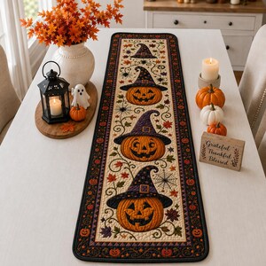 May include: A rectangular table runner featuring a cream-colored center panel with three orange jack-o'-lanterns wearing purple witch hats, surrounded by swirling vines, spiderwebs, and autumn leaves, all framed by a dark border with small pumpkins.