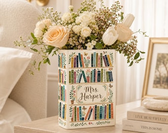 May include: A rectangular white ceramic vase featuring a colorful illustration of a bookshelf filled with various books and potted plants. The center of the vase displays the text Mrs. Harper in a decorative script font, surrounded by floral accents.