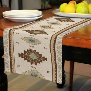 May include: Cream-colored table runner with a geometric pattern in brown, teal, and gold. The runner features a repeating diamond and cross design, bordered by a decorative edge. The runner is placed on a dark wooden table, with white plates and a fruit bowl visible.