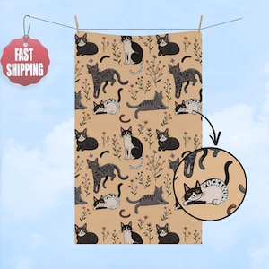 May include: Tan-colored towel with a repeating pattern of various cat illustrations, flowers, and crescent shapes. The cats are in different poses and colors, including black, gray, and white. The towel is hanging on a clothesline.