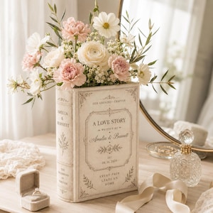May include: A cream-colored book-shaped vase featuring the text A LOVE STORY, WRITTEN BY Amelia & Bennett, EST. 08.16.2026, OUR GREATEST CHAPTER, and EVERY PAGE LEADS US HOME, filled with a bouquet of white and pink flowers and green foliage.