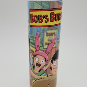 May include: A tall, cylindrical tumbler featuring characters from the animated series "Bob's Burgers." The tumbler has a colorful design with the show's logo in red and yellow. The characters Louise Belcher and Gene Belcher are prominently displayed.