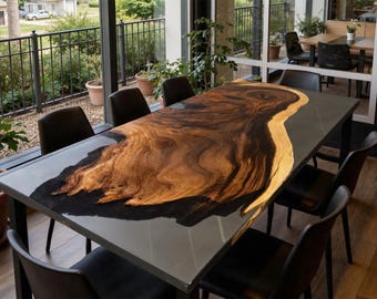 Può includere: Un grande tavolo da pranzo rettangolare con un design unico a venature di legno. Il tavolo presenta una superficie scura e lucida con un motivo in legno naturale nei toni del marrone e dell'oro. È sostenuto da una struttura in metallo scuro e circondato da sedie nere. Il tavolo misura circa 240 cm di lunghezza.