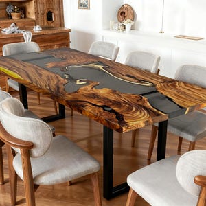 May include: A large, rectangular dining table with a live edge wooden top and a dark grey epoxy resin centre. The table has black metal legs. Eight grey upholstered chairs with wooden frames surround the table.