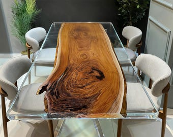 Op de afbeelding: Een moderne eettafel met een houten blad met een natuurlijke rand, ingekapseld in helder acryl. De tafel wordt omringd door vier grijs gestoffeerde stoelen met houten poten. Het houten blad heeft een rijke bruine kleur.