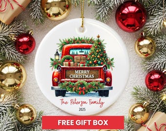 May include: A white ceramic Christmas ornament featuring a red truck carrying a Christmas tree and gifts. The ornament has the text "Merry Christmas" and "The Peterson Family 2025". Surrounded by red and gold Christmas ornaments and evergreen branches.