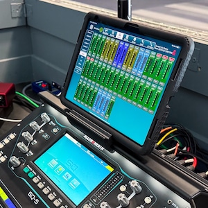 Puede incluir: Una consola de mezcla de audio digital con una tableta adjunta que muestra una interfaz de mezcla. La consola es negra con numerosas perillas, botones y una pantalla. También se ve una interfaz de audio roja y otros equipos.