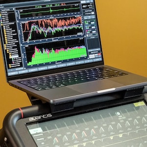 Peut inclure: Un ordinateur portable gris foncé est posé sur un support noir au-dessus d'une console de mixage audio Avantis. L'écran affiche un logiciel d'analyse audio complexe avec plusieurs graphiques de fréquences colorés et des données.