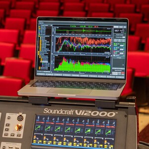 May include: A silver laptop displaying complex audio analysis software with colourful frequency graphs and data charts, mounted on a black Soundcraft Vi2000 digital mixing console featuring illuminated control panels and various input channels.