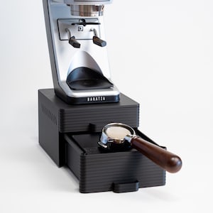 May include: A silver and black Baratza coffee grinder with a wooden-handled portafilter in the drawer. The grinder sits on a black base with a drawer. The Baratza logo is visible.