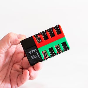 May include: A black Sandisk memory card adapter with red and green detailing. The adapter, held in a hand, features multiple slots for various memory card types. It's designed for memory card format conversion.