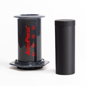 Puede incluir: Cafetera AeroPress, con una cámara cilíndrica gris oscuro con texto "AeroPress" rojo y marcas de medición. También hay un cilindro negro separado, sobre un fondo blanco.