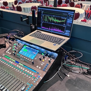 May include: An Allen & Heath SQ-5 digital audio mixing console with a silver laptop resting on top. The laptop screen displays real-time audio frequency analysis software with green, red, and purple waveforms. Black cables are connected to the console.
