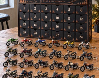 May include: A black advent calendar box labelled MOTO TRAILS 24 MINI MOTO KEYRINGS with 24 numbered drawers. In front are 24 miniature motorcycle keychains in various colours including green, red, blue, orange, yellow, white, and black.