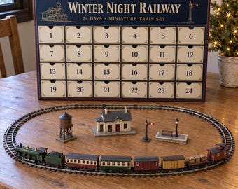 May include: A Winter Night Railway 24-day miniature train set advent calendar box with numbered drawers. Below the box sits an oval train track with a green locomotive, various train cars, a water tower, a station building, and a platform.