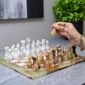 May include: A marble chess set with white and brown pieces on a green and white board. A hand is holding a brown chess piece above the board. The chess board is on a marble surface.