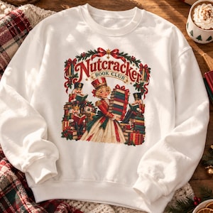 May include: A white crewneck sweatshirt featuring a colorful graphic print of a Nutcracker Book Club scene. The design includes a central figure in a red uniform holding a stack of books, surrounded by nutcracker characters, greenery, and red bows.