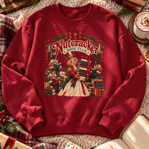 May include: A dark red crewneck sweatshirt featuring a graphic print of a Nutcracker Book Club scene. The design shows a central figure holding a stack of books surrounded by nutcracker characters, festive greenery, and ribbons in a holiday style.