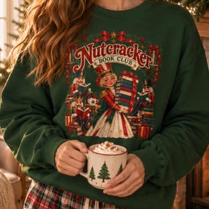 May include: A dark green crewneck sweatshirt featuring a graphic print of a Nutcracker Book Club scene with a central figure holding a stack of books, surrounded by nutcracker characters, festive greenery, and the text Nutcracker Book Club.