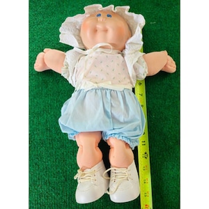 May include: A vintage Cabbage Patch Kid doll, featuring a soft, peach-colored face and limbs, blue eyes, and a sweet expression. The doll is dressed in a light blue outfit with a floral print top, matching bloomers, and white shoes. A white bonnet completes the look.
