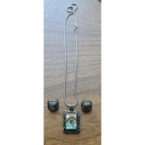 Puede incluir: Un conjunto de joyas plateadas con un collar y pendientes a juego. El collar tiene un colgante rectangular con un diseño de concha de abulón. Los pendientes tienen forma cuadrada con acentos similares de concha de abulón. El conjunto se muestra sobre una superficie de madera.