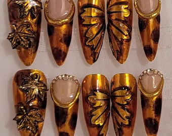 May include: Set of ten artificial nails with a fall theme. The nails feature a glossy orange and brown color scheme with leaf and jewel accents. Some nails have a leopard print design.