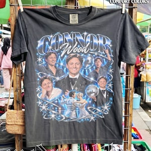 May include: A charcoal gray short-sleeve t-shirt featuring a graphic print of Connor Wood. The design includes multiple photos of the person, a disco ball, and star accents. The text "CONNOR" and "Wood" are printed in a bold, stylized font.