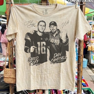Op de afbeelding: Een beige t-shirt met korte mouwen en een zwart-witte afbeelding van Jared Goff en Dan Campbell. De print bevat de tekst "Jared Goff" en "Dan Campbell" met handtekeningen. Het shirt heeft een ronde hals en een Comfort Colors merklabel.