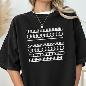 May include: Black t-shirt with a white graphic design featuring various sewing stitch patterns. The shirt has a relaxed fit and short sleeves. The design includes straight stitches, wavy lines, and a cross-stitch pattern.